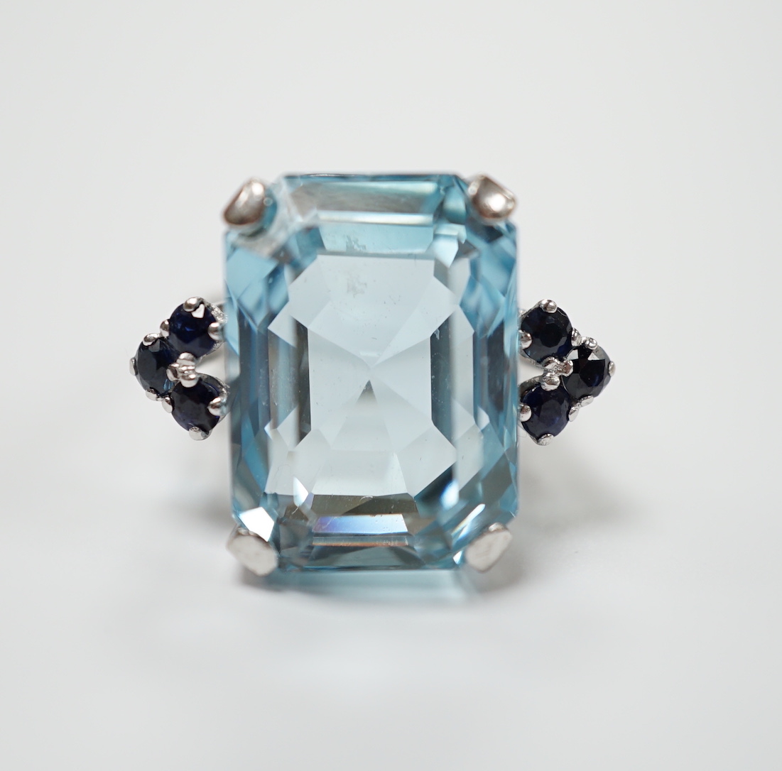 An 18ct white metal and single stone emerald cut aquamarine set dress ring, with six stone sapphire set shoulders, size L, gross weight 11.8 grams.